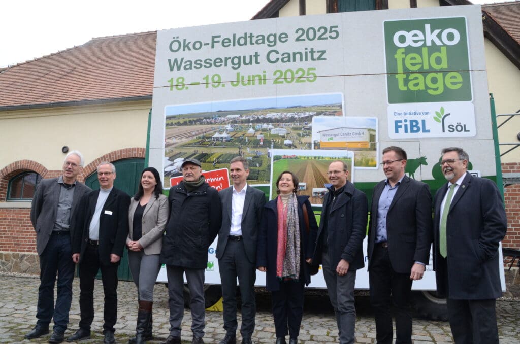 Personen vor einem Hinweisschild. das auf die Öko-Feldtage 2025 auf dem Wassergut Canitz verweist.